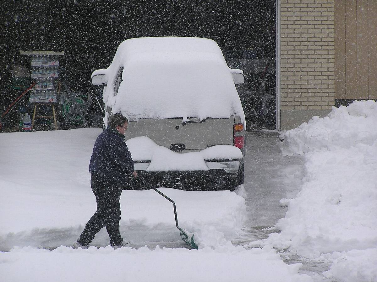 A person shoveling a snow-covered driveway beside a buried pickup truck while snow is still falling.